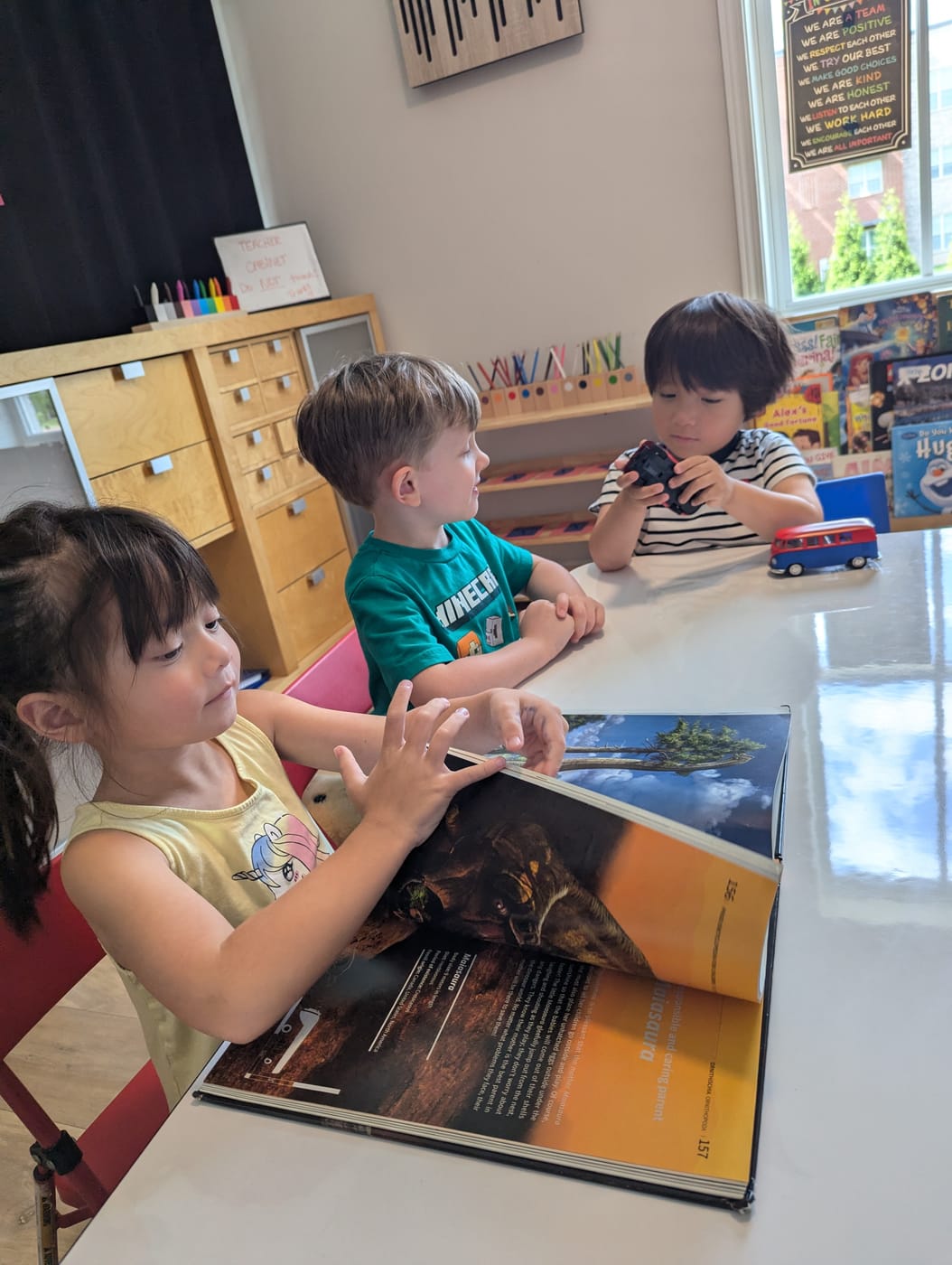 Children reading a dinosaur book and playing with toy cars at a table